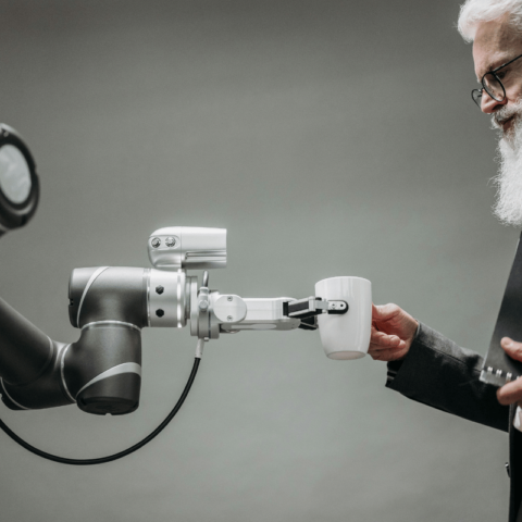 Elderly man with a beard using a robotic arm to hold a cup, demonstrating human-robot interaction in a modern setting.