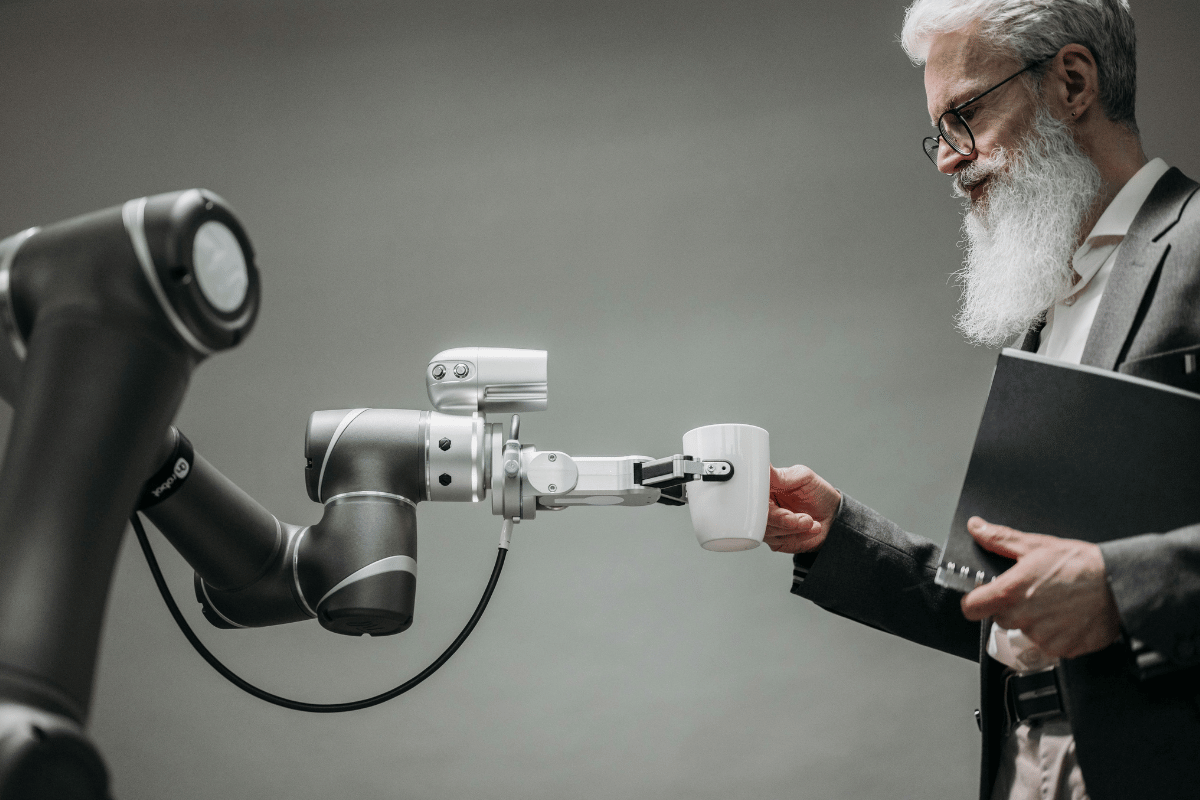 Elderly man with a beard using a robotic arm to hold a cup, demonstrating human-robot interaction in a modern setting.