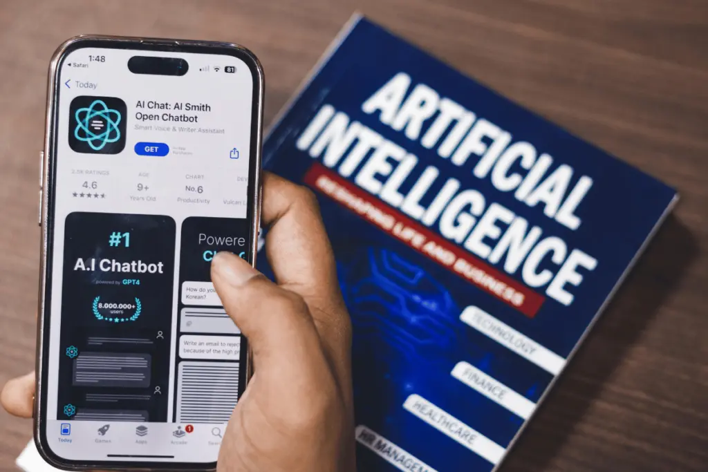 Person holding a smartphone displaying an AI chatbot app, with an Artificial Intelligence book in the background.