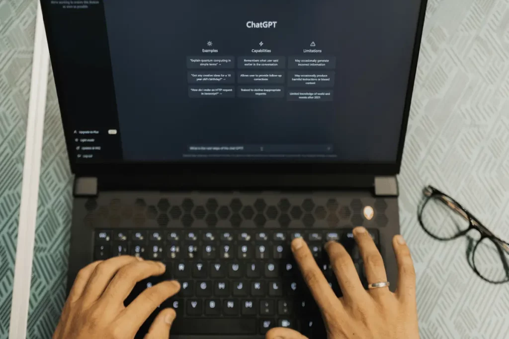 Hands typing on a laptop with ChatGPT interface on screen, showing examples, capabilities, and limitations in a user-friendly design.