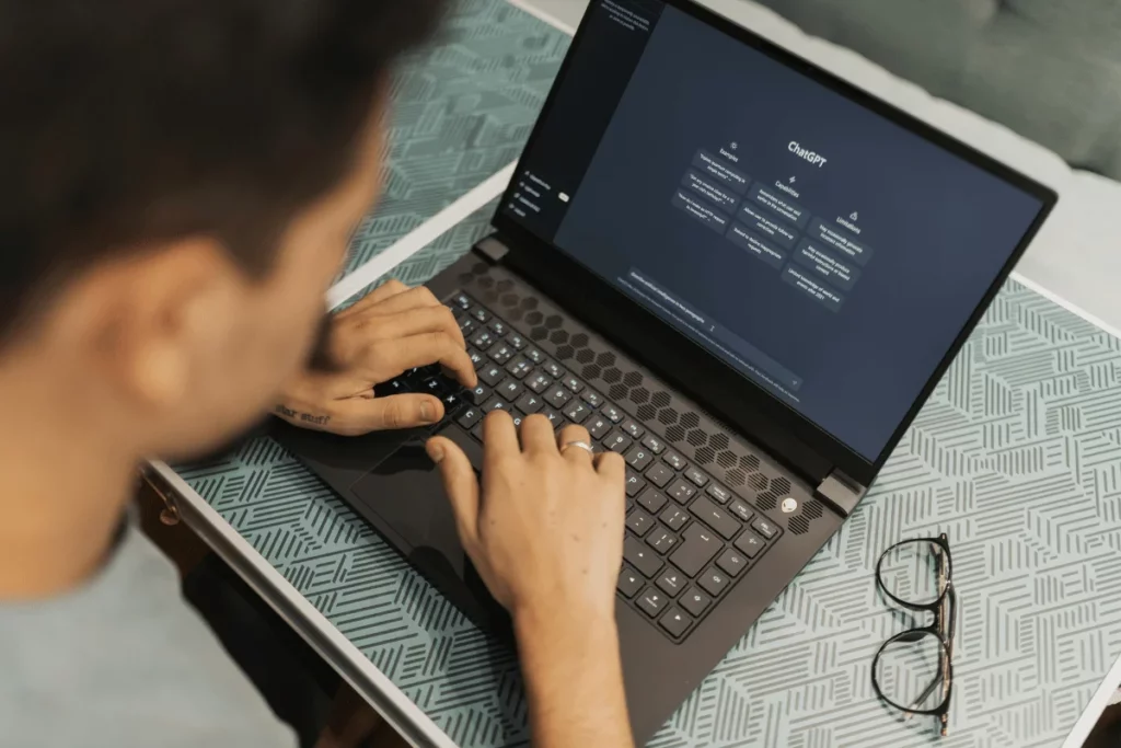 Man typing on a laptop, using ChatGPT, with a pair of glasses on the table.