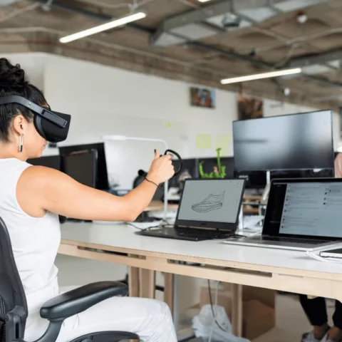 Woman wearing VR headset while designing a 3D shoe model at her workstation in an office.