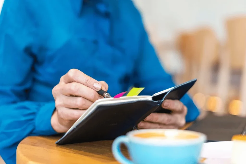 A person writing in a notebook in a cafe with a cup of coffee.