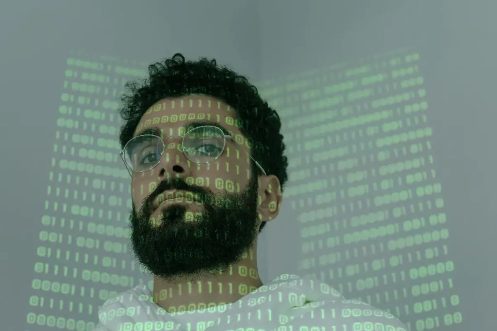 Man with glasses and beard, green binary code projected on his face, looking up.