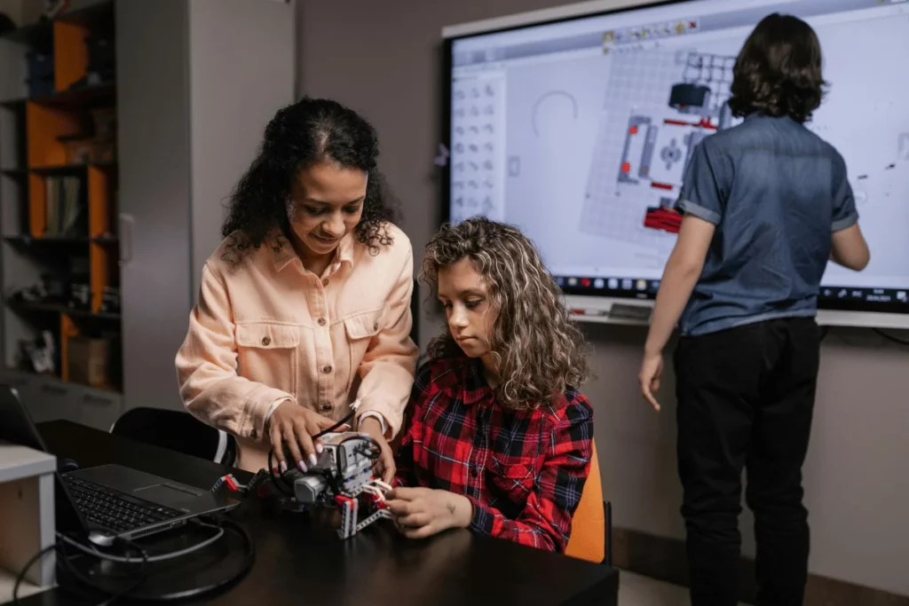 Teacher guiding a student in building a robot, with another student working on a smartboard in the background.