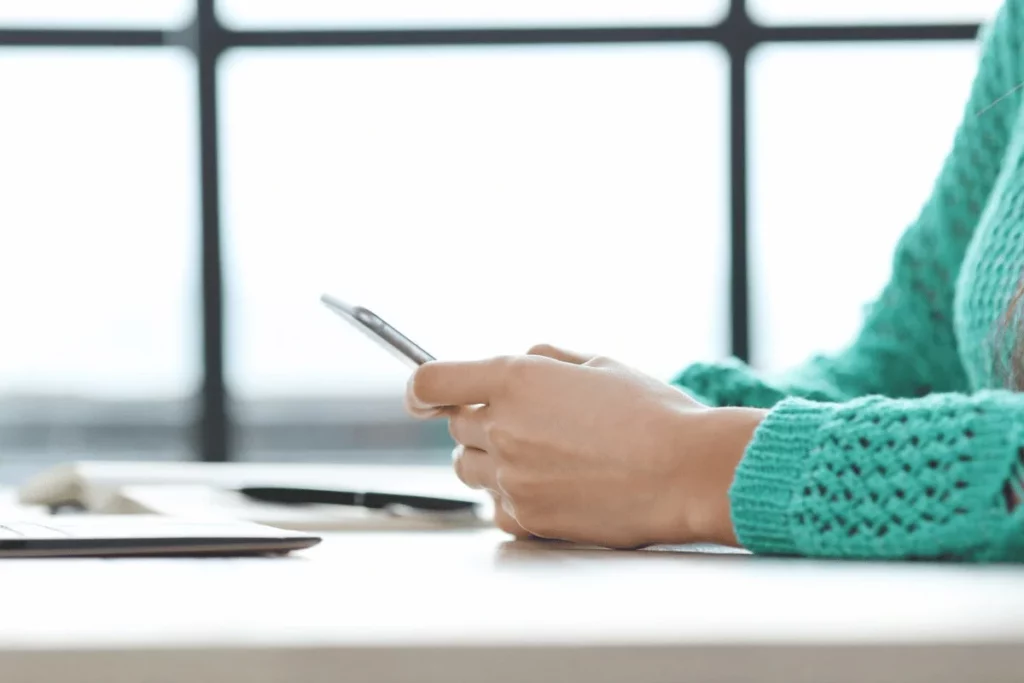 A person uses a cell phone, with a laptop in front while sitting at a desk.