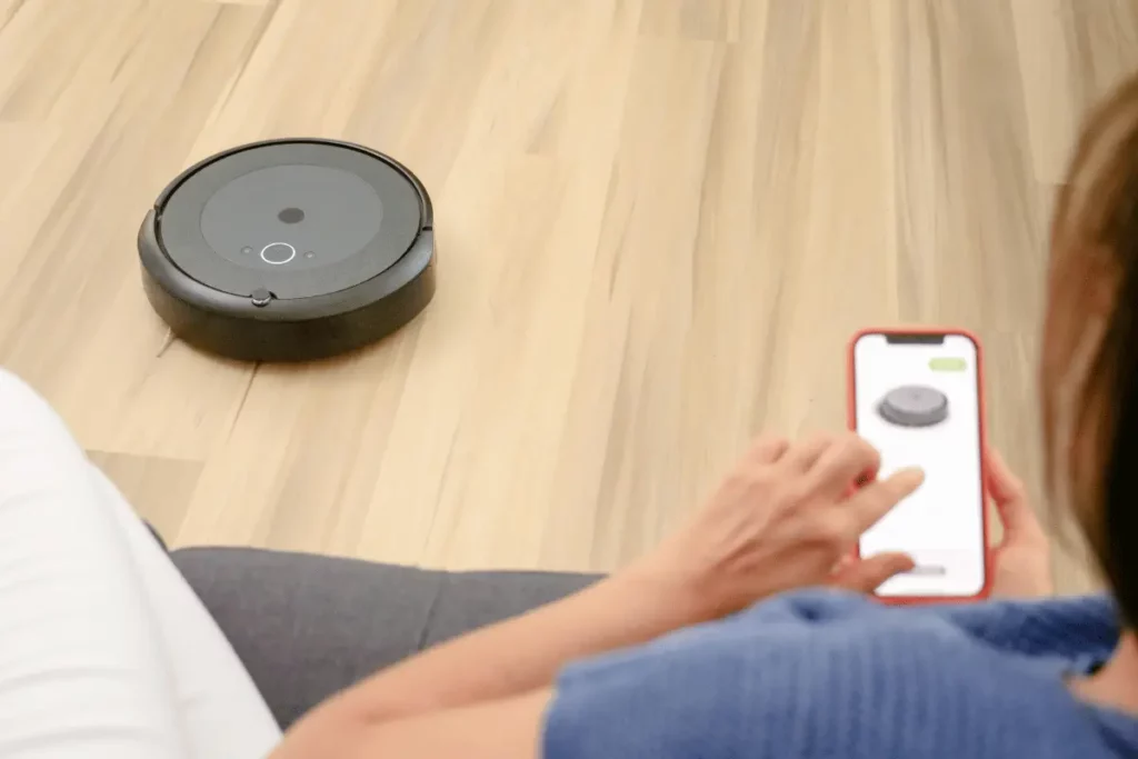 Person using a smartphone app to control a robotic vacuum cleaner on a wooden floor, highlighting smart home technology.
