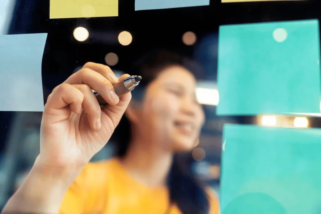 Close-up of a hand holding a pen, writing on a glass board with colorful sticky notes.