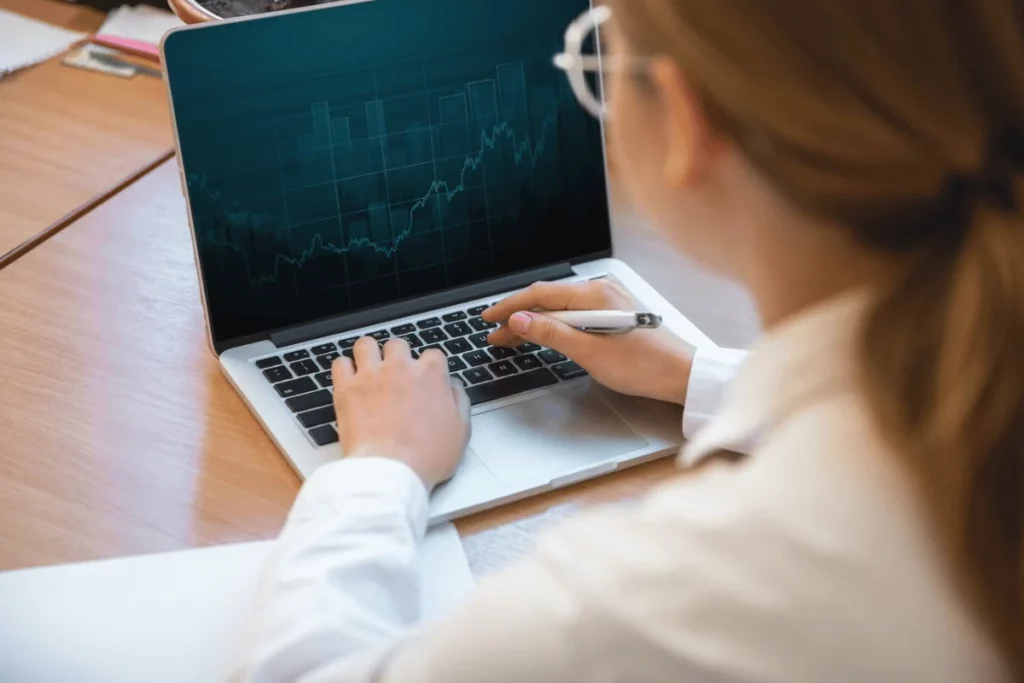 Female reviewing stock market trends on a laptop, taking notes with a pen.