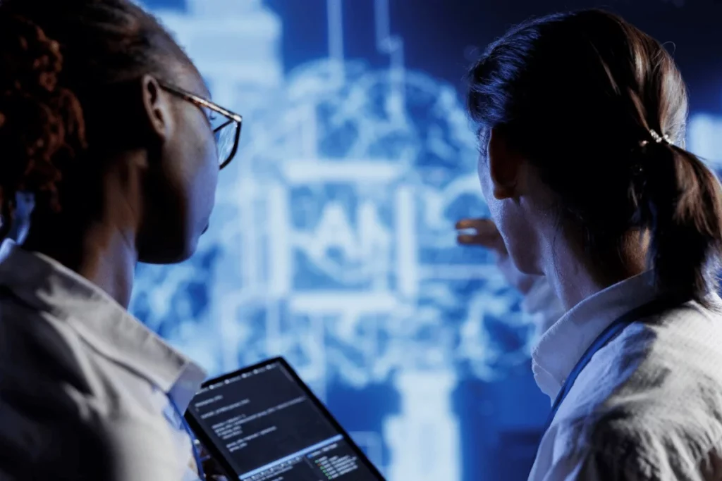 Two professionals analyzing data on a large screen, with one holding a tablet displaying code.