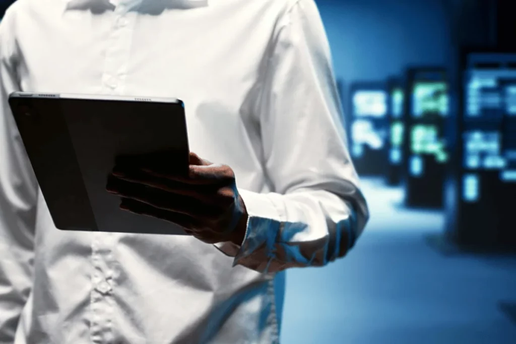 Professional holding a tablet in a data center with blurred server racks in the background.