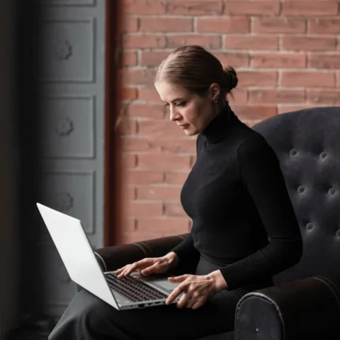 Woman in a black turtleneck sitting in a chair working on a laptop near a large window.