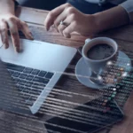 Person working on a laptop at a wooden desk with a cup of coffee and visible code overlay.