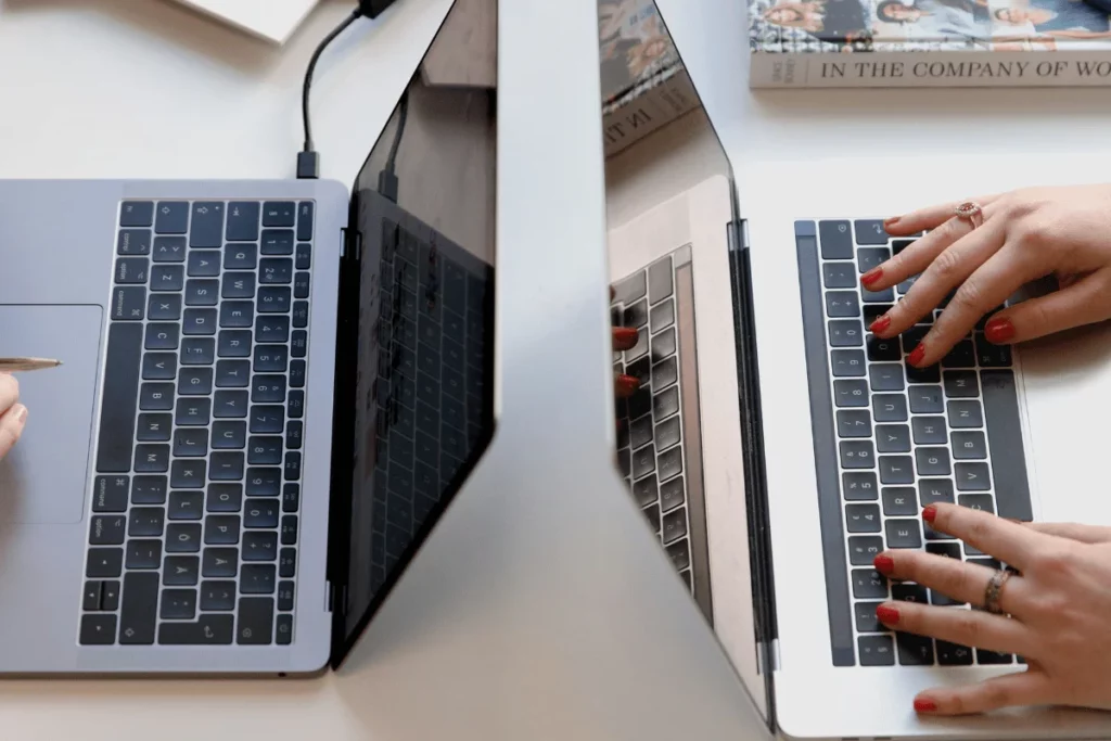 Two laptops side by side with people typing on their keyboards.