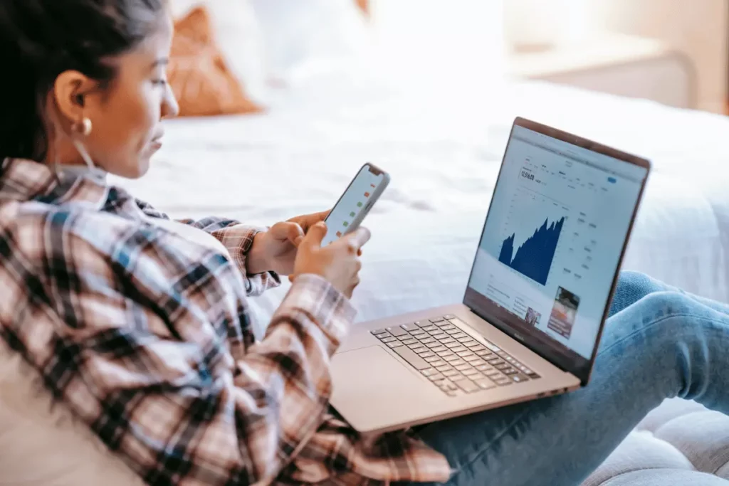 Person analyzing stock market graphs on a laptop and mobile phone, relaxing on a bed.