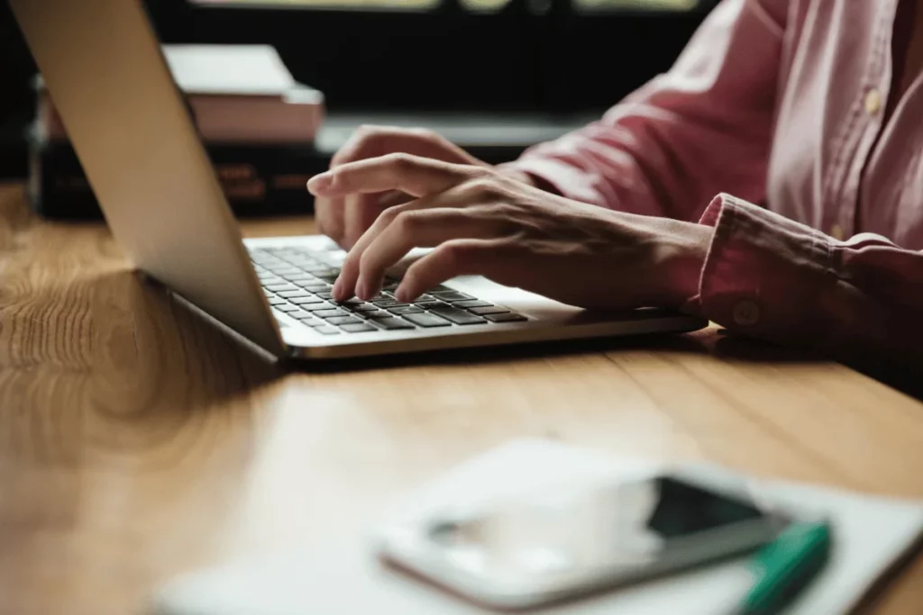 Hands typing on a laptop keyboard in a cozy, book-filled workspace with a smartphone and notebook in the foreground.