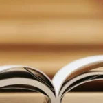 Open book on a wooden table with a black travel mug and stacked books in the background.