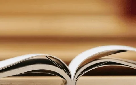 Open book on a wooden table with a black travel mug and stacked books in the background.