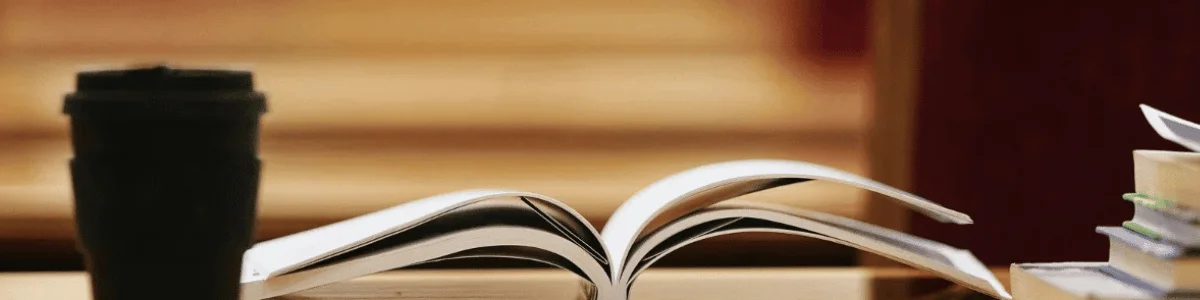 Open book on a wooden table with a black travel mug and stacked books in the background.