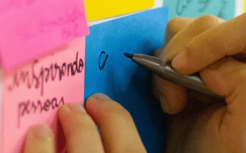 Close-up of a hand writing on a blue sticky note among other colorful notes on a board.