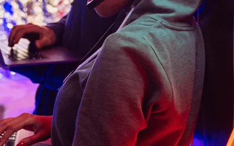 A person in a hoodie types on a laptop, illuminated by colorful neon lights, while another individual stands behind with a laptop.