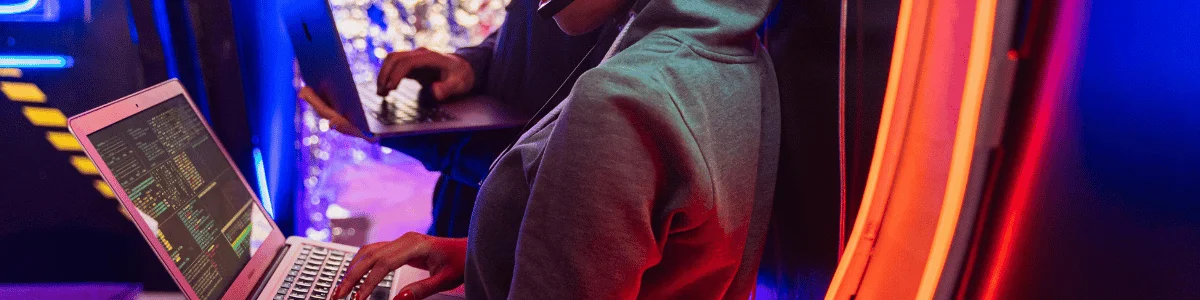 A person in a hoodie types on a laptop, illuminated by colorful neon lights, while another individual stands behind with a laptop.