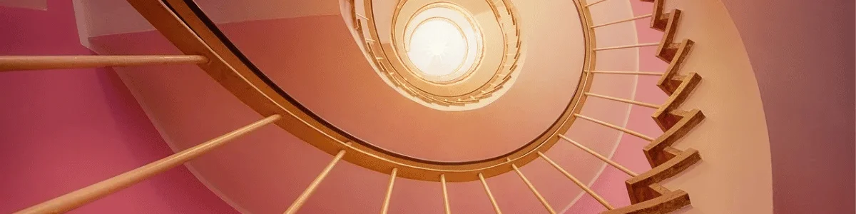 Looking up at a spiral staircase with beige railings and pink walls, illuminated by a bright light at the top.