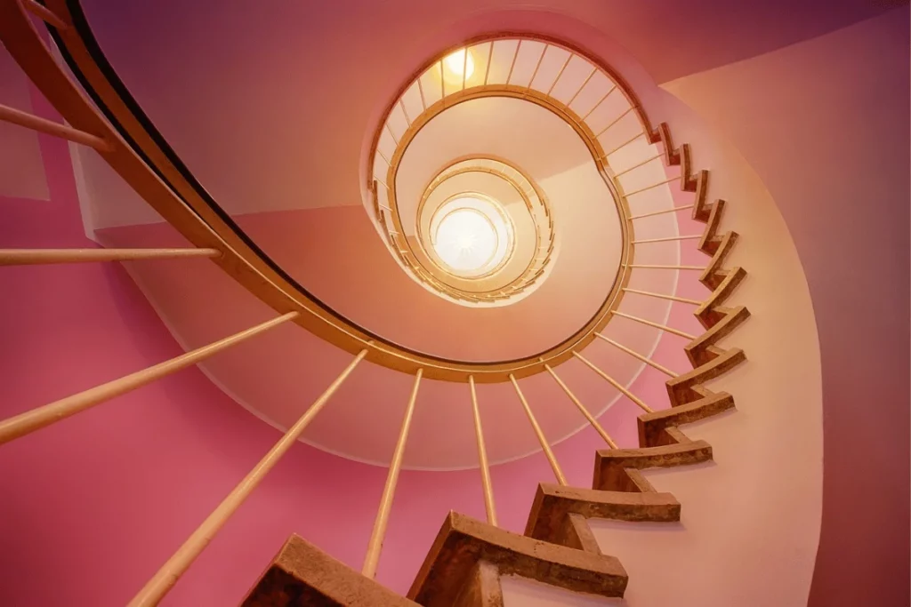 A spiral staircase viewed from above, with warm lighting and pink walls creating an inviting, modern aesthetic.