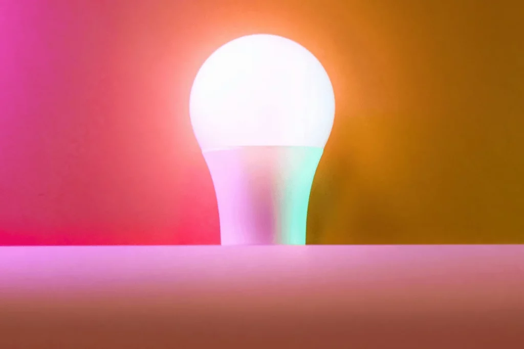 A close-up of a colorful light bulb glowing with shades of pink, purple, and green against an orange backdrop.