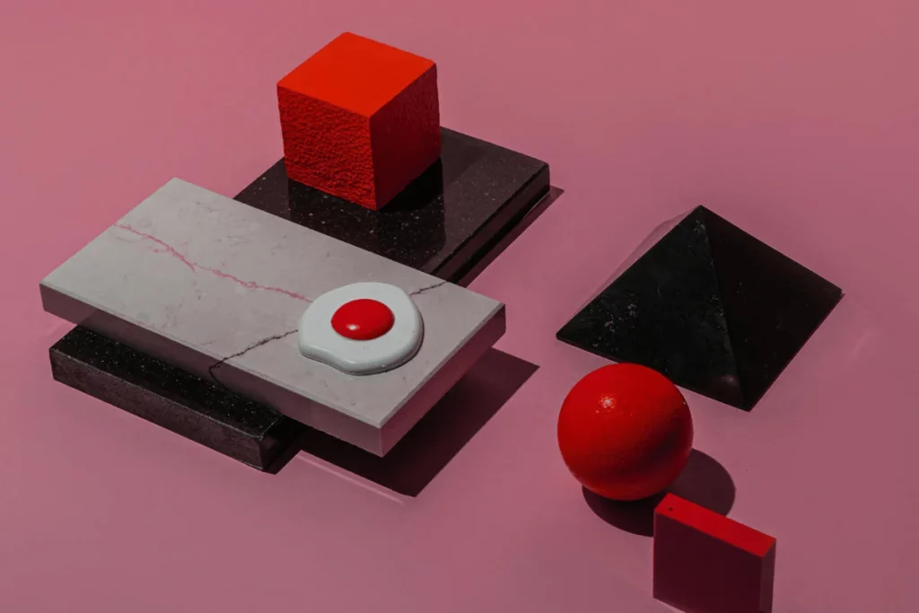 A collection of colorful geometric shapes, including red spheres, cubes, and pyramids, set against a pink background.
